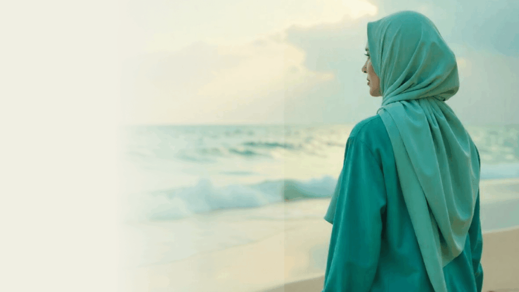 Muslim woman in teal hijab looking over the sea at sunrise, symbolising calm after healing from trauma